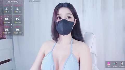 Media: Video of an East Asian woman with long dark hair, wearing a black face mask and light blue halter top, seated indoors with white curtains and a calendar screen overlay.