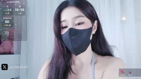 Media: A video of a young East Asian woman with long, straight black hair, wearing a black face mask and a light gray top, indoors, with a video overlay showing her live stream.