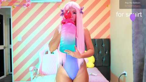 Video of a young woman with long, pastel pink hair, wearing a blue one-piece swimsuit, drinking a pink beverage, standing in a pastel-striped bedroom with a tufted black headboard.