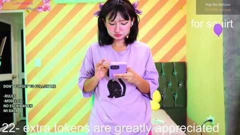 A young Asian woman with straight black hair, wearing a purple sweater with a black cat graphic, holds a smartphone, in a colorful room with striped wallpaper and a green headboard.