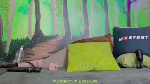 Media: Video of a bedroom with a green forest mural, a yellow pillow, a dark pillow with \"mrstbet\" text, a bottle, and a rolled-up blanket.