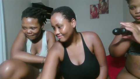 Media: Video of two women with dark skin, one with dreadlocks in a tank top, the other in a black tank top, sitting on a red couch, smiling. Background includes a white wall with red artwork.