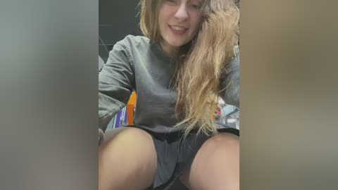 Media: A video of a young woman with long, wavy hair, wearing a gray sweatshirt and shorts, sitting with legs apart, smiling. The background is blurry and neutral-colored.