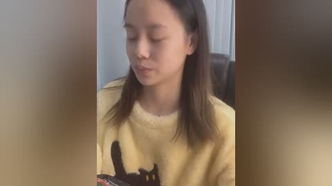 Media: Video of a young Asian girl with straight black hair, wearing a yellow sweatshirt with a cat graphic, sitting indoors, eyes closed, serene expression.