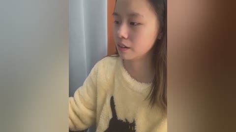 Media: A video of a young Asian girl with straight black hair, wearing a yellow fleece sweater, looking slightly to the side. The background features a blurred wooden door and a wall.