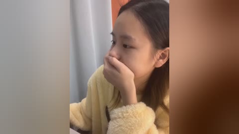 Media: Video of a young Asian girl with straight black hair, wearing a yellow fleece robe, resting her chin on her hand, looking introspective. Background is a plain, off-white wall with a hint of orange.