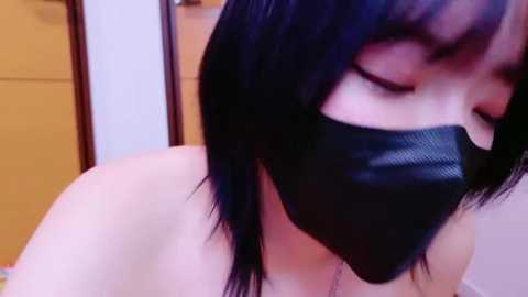 Media: Video of an Asian woman with shoulder-length black hair and fair skin, wearing a black face mask, leaning forward slightly, in a dimly lit room with wooden furniture and a blurred background.