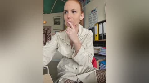 Media: Video of a pale-skinned woman with red hair, wearing a beige trench coat and black fishnet stockings, sitting in an office, looking contemplative.