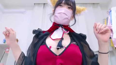 Video of a young woman with fair skin, large breasts, wearing a revealing red and black lace lingerie set, black cape, and cat ears, indoors with white walls and hanging clothes.