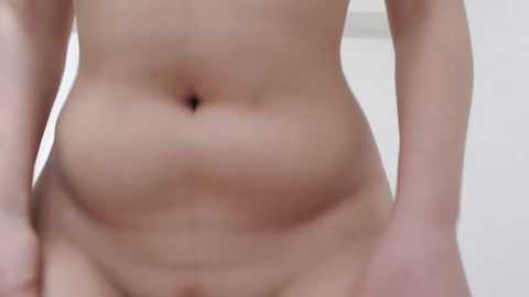 Media: Video of a nude, light-skinned person with a slightly protruding belly, wearing no clothing, standing indoors with a blurred white background.