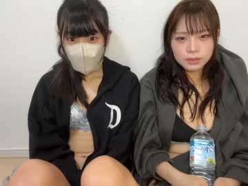 Media: Video of two young East Asian women, wearing masks, sitting on a light-colored floor. One wears a black hoodie with a white logo, and the other a green jacket over a black bra. They hold water bottles, against a plain white background.