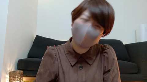 Video of a person with short, brown hair, wearing a brown, button-up shirt and a gray mask, sitting on a black couch in a modern, minimalistic living room.