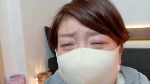 Media: A close-up video of a young Asian woman with short, brown hair, wearing a white face mask, looking tired and slightly sad. The background is a blurred indoor setting with a wooden table and a white wall.