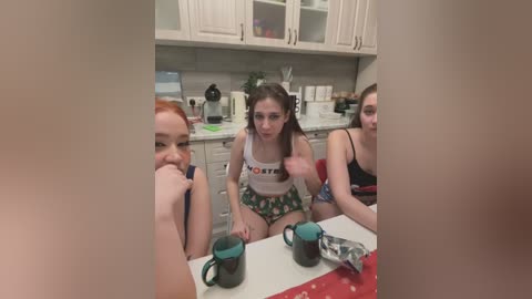 Media: Video of three young women in casual, revealing outfits sitting at a kitchen counter. One woman in a Hooters tank top and shorts gestures with a playful expression.