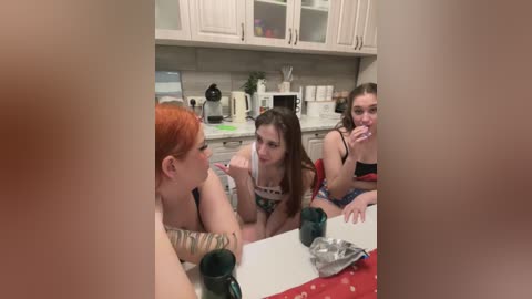 Media: Video of three young women in a kitchen, one with red hair, one with dark hair, and one with long brown hair. They are seated at a table, drinking from green mugs.