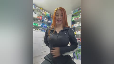 Video of a curvaceous woman with long, straight red hair, wearing a black and white Adidas track jacket, standing in a brightly lit, organized pharmacy with colorful medicine shelves in the background.