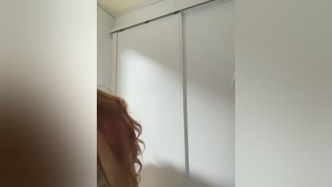 Video of a woman with long, curly brown hair partially visible in a narrow, dimly lit hallway with white walls and a sliding glass door to the right.