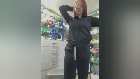 Video of a red-haired woman with fair skin, wearing a black athletic jacket and pants, standing in a brightly lit pharmacy with colorful products in the background.