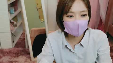 Media: A video of an East Asian woman with light skin and short brown hair, wearing a lavender face mask and light blue button-up shirt, sitting in a pink carpeted room with a mirror, pink and green walls, and a yellow towel hanging.