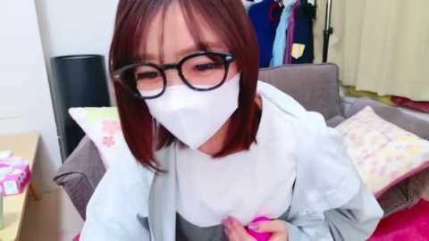 Media: Video of an Asian woman with straight, shoulder-length brown hair, wearing black-framed glasses, a white mask, and a light blue blouse. She is seated on a brown couch in a cluttered room with clothes hanging in the background.