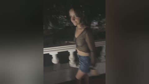 Media: A video of a woman with dark hair, wearing a sheer top and denim shorts, walking on a balcony at night. The background is dark and blurry, with a railing and trees visible.