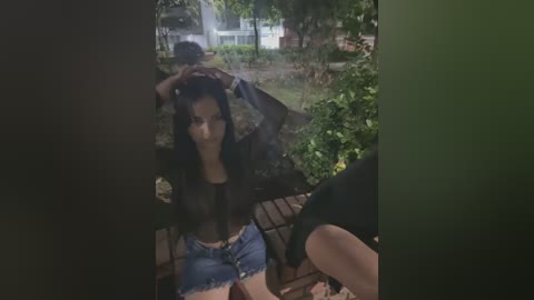 Media: Video of a young woman with long black hair, wearing a black sheer top and denim shorts, sitting on a wooden bench in a lush garden.