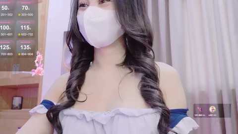 Media: Video of an Asian woman with long black hair and a white face mask, wearing a strapless, off-shoulder white dress. She stands indoors against a wooden backdrop with a digital display showing temperature and humidity.