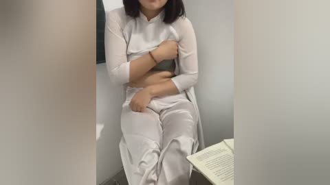 Media: Video of a woman in a white, semi-transparent dress sitting on a chair in a bare, dimly lit room. She covers her chest with her left arm. An open book lies on a table beside her.