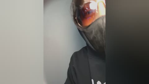 Media: A close-up video of a person wearing a black hoodie and a black face mask with a reflective visor, partially obscured by a blurred background.