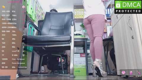 Media: Video of a woman in maroon pants and heels standing behind a desk in a cluttered office. Background shows colorful filing cabinets and a black leather chair.