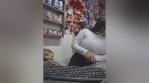 Media: A video of a woman in a white long-sleeve shirt, sitting at a desk, with a keyboard and various snacks on shelves in the background.