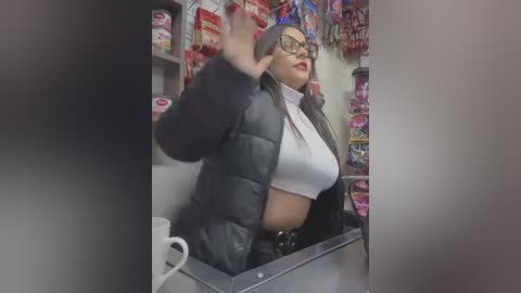 Media: Video of a Latina woman with long dark hair, wearing glasses, a black puffer jacket, and a white crop top, standing in a candy store with colorful displays of candy in the background.