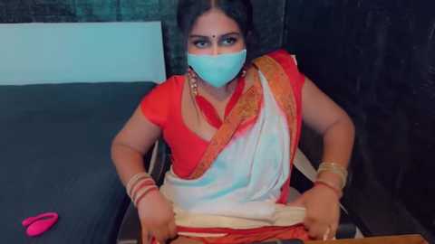 Media: Video of an Indian woman in a red sari with a white border, wearing a blue face mask, seated on a dark bench. Background includes a gray wall and a pink object on the left.