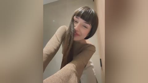 Media: Video of a young Asian woman with straight, shoulder-length brown hair, wearing a beige cardigan, bending over to take a selfie. She's smiling slightly. The background features beige walls and a mirror.