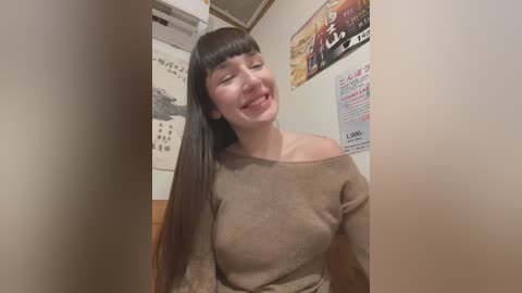 Media: Video of a smiling woman with long black hair, wearing a beige off-shoulder sweater, standing in a narrow hallway with Japanese posters on the walls.