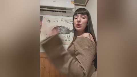 Media: A video of an Asian woman with long, straight black hair and fair skin, wearing a beige sweater, leaning against a calendar and air conditioner in a dimly lit room.