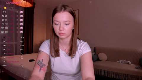 Media: Video of a young Caucasian woman with straight, shoulder-length brown hair, wearing a white t-shirt. She has a small tattoo on her left arm. Background shows a dimly lit bedroom with a bed, curtains, and a TV displaying a live feed.