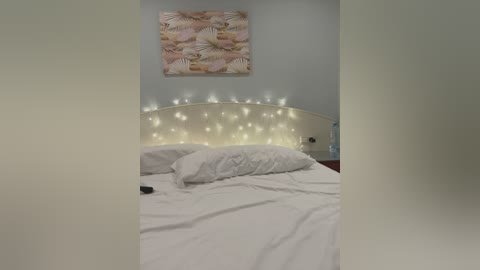 Media: Video of a neatly made bed with white linens, surrounded by a string of fairy lights on a beige headboard. A floral painting hangs on the light gray wall, with a subtle pink and white color scheme.