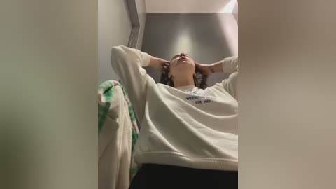 Media: Video of a person with short hair, wearing a white sweatshirt, lying on their back with hands behind their head, inside a narrow, dimly lit space with white walls.