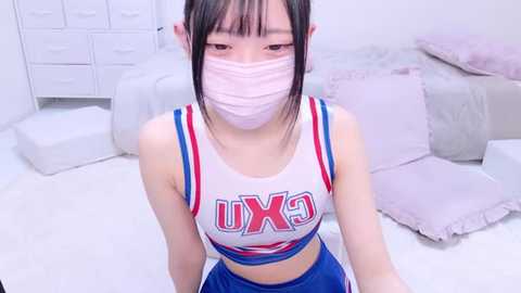 Media: A video of an East Asian woman with straight black hair and fair skin wearing a white crop top with \"UXK\" in red and blue letters, blue shorts, and a pink face mask. She sits on a white floor with a bed and white drawers in the background.