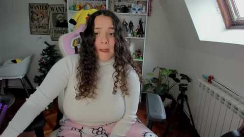 Media: Video of a curvy, light-skinned woman with curly brown hair, wearing a tight white top and pink pajama pants, seated in a cluttered, brightly lit room with a Christmas tree and bookshelves.