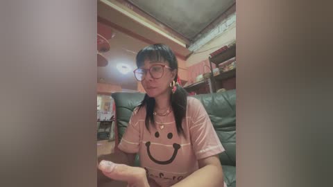 Media: Video of an Asian woman with long black hair, wearing glasses, pink T-shirt with a smiling face, sitting on a green leather couch in a cluttered room.