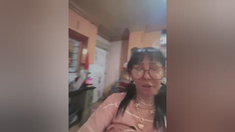 Media: A video shows an older woman with glasses, wearing a pink blouse, in a domestic setting with cabinets and a door in the background.