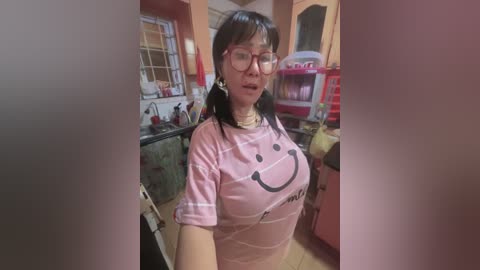 Media: Video of an Asian woman with glasses, large breasts, wearing a pink \"Kawaii\" T-shirt, standing in a cluttered kitchen with pink and orange walls, a sink, and a toy vending machine in the background.