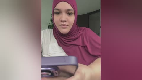 Video of a young woman with fair skin and light brown hair wearing a maroon hijab and white shirt, focused on a smartphone in her hands. Background features a blurred indoor setting with neutral tones.