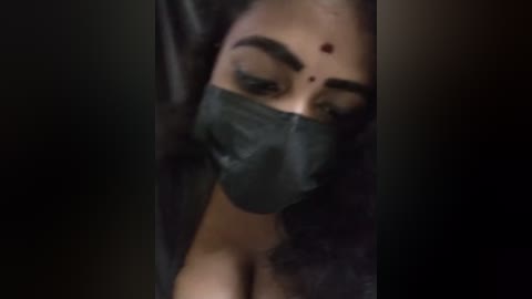 Media: A video of a close-up of a woman with medium skin tone, wearing a black face mask, with thick, dark eyebrows, and a small red bindi on her forehead.