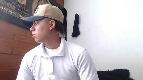 Media: Video of a young man with light skin, wearing a tan baseball cap, white polo shirt, and ear studs, standing against a white wall with a black jacket and brick wall in the background.