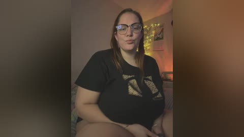 Media: A video of a plus-sized, light-skinned woman with glasses, long brown hair, and tattoos, wearing a black t-shirt and sitting indoors, with a warm, dimly-lit background.