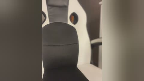 Media: Video of a modern, minimalist office chair with black upholstery and a white backrest, viewed from the front, partially blurred. The chair sits on a beige carpet, with a gray wall and dark wood desk in the background.