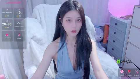 Media: A video of a young East Asian woman with long black hair, wearing a light blue halter top, sitting on a white bed in a soft, pink-lit bedroom.